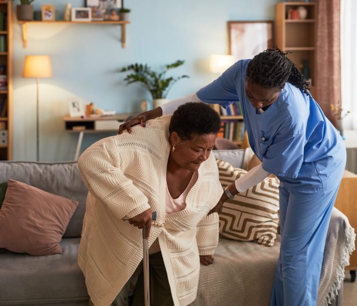 Senior Black woman using cane receiving assistance from young adult Black female nurse in scrubs helping her stand up from sofa in living room setting during home care visit